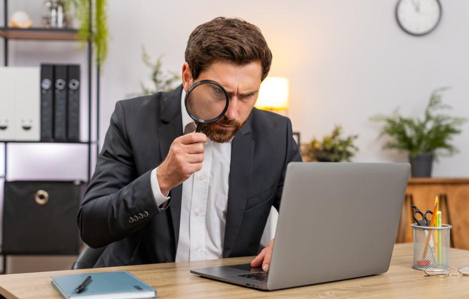  Um homem de terno usa uma lupa para analisar minuciosamente as informações na tela de um notebook.