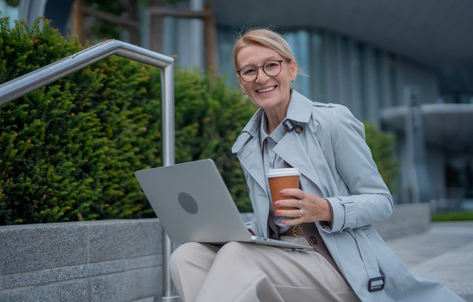 Mulher jovem e sorridente sentada ao ar livre com um laptop e um café, desfrutando do ambiente urbano e do clima agradável, transmitindo produtividade e bem-estar.
