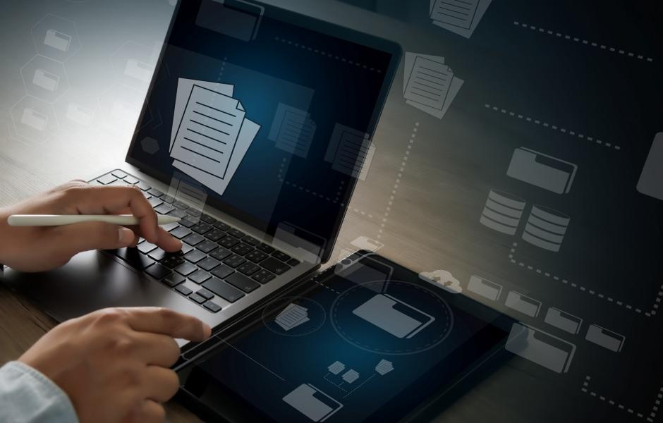 Mãos operando um notebook e segurando uma caneta digital ao lado de um tablet. Telas e fundo exibem elementos gráficos transparentes conectados, representando gerenciamento de arquivos, pastas virtuais e bancos de dados.