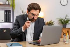  Um homem de terno usa uma lupa para analisar minuciosamente as informações na tela de um notebook.