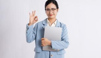 Mulher profissional de negócios com óculos, cabelo escuro, vestida formalmente com terno azul claro, sorrindo e fazendo sinal de aprovação com a mão, segurando um laptop e transmitindo confiança e otimismo.