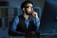 Homem barbudo de óculos e colete trabalhando concentrado em frente a um computador em um escritório escuro à noite.