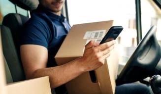 Entregador sentado no banco do motorista de uma van de entregas, segurando uma caixa de papelão com código de barras e verificando o celular. Ao fundo, outras caixas e o painel do veículo.