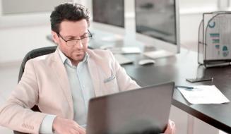 Homem de terno claro e óculos trabalhando focado em seu notebook em um escritório bem iluminado. A imagem transmite profissionalismo, seriedade e dedicação à gestão empresarial.