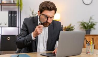  Um homem de terno usa uma lupa para analisar minuciosamente as informações na tela de um notebook.