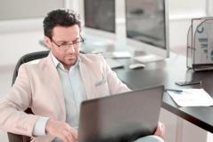 Homem de terno claro e óculos trabalhando focado em seu notebook em um escritório bem iluminado. A imagem transmite profissionalismo, seriedade e dedicação à gestão empresarial.