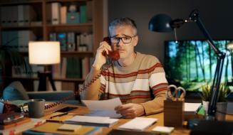 Homem de óculos com cabelo grisalho, usando camisa listrada, trabalha em escritório com computador, livros e cadeiras, enquanto conversa ao telefone de casa.