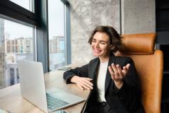 Mulher jovem e sorridente com cabelo curto em uma videochamada no laptop. Ela gesticula com a mão, veste blazer preto e está sentada em um escritório moderno com uma grande janela ao fundo.