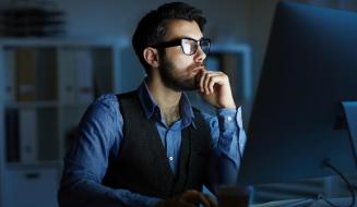 Homem barbudo de óculos e colete trabalhando concentrado em frente a um computador em um escritório escuro à noite.