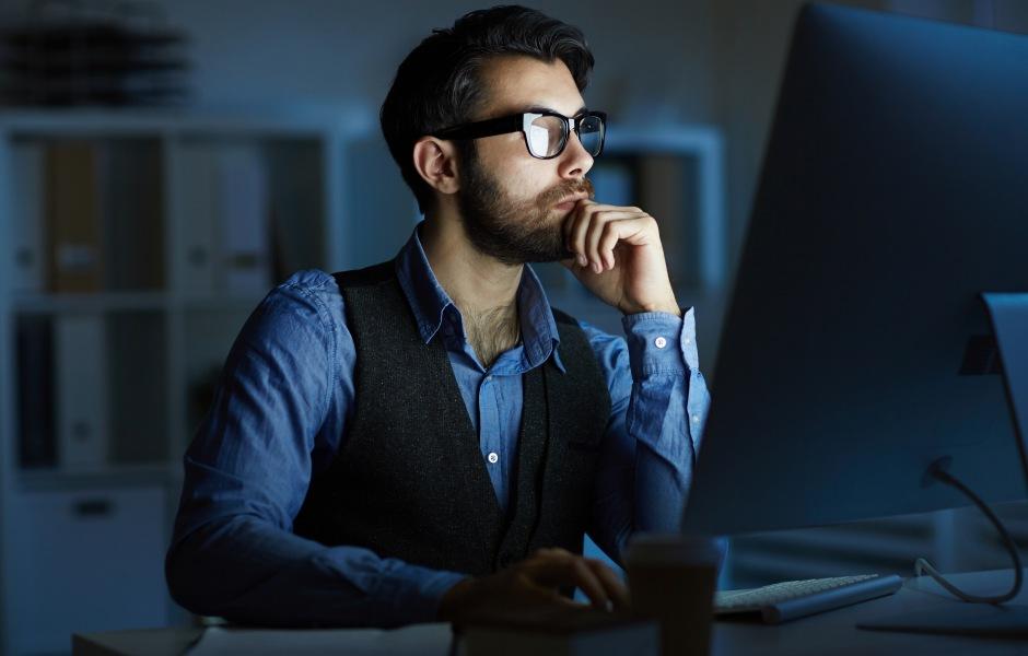 Homem barbudo de óculos e colete trabalhando concentrado em frente a um computador em um escritório escuro à noite.