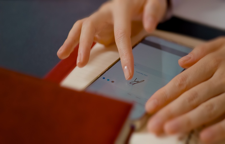 Pessoa usando smartphone para fazer assinatura digital, sentado em mesa com documentos ao redor, rotina de trabalho ou assinatura digital.