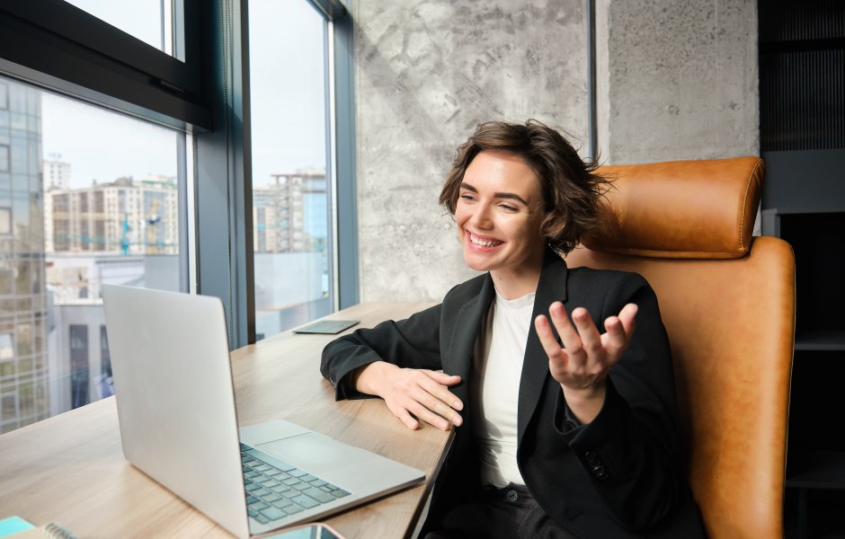 Mulher jovem e sorridente com cabelo curto em uma videochamada no laptop. Ela gesticula com a mão, veste blazer preto e está sentada em um escritório moderno com uma grande janela ao fundo.