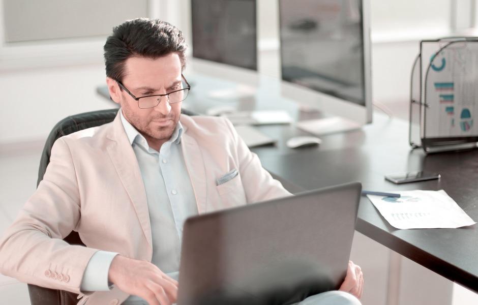 Homem de terno claro e óculos trabalhando focado em seu notebook em um escritório bem iluminado. A imagem transmite profissionalismo, seriedade e dedicação à gestão empresarial.
