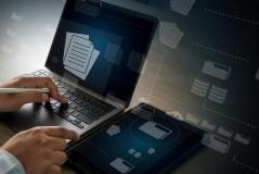 Mãos operando um notebook e segurando uma caneta digital ao lado de um tablet. Telas e fundo exibem elementos gráficos transparentes conectados, representando gerenciamento de arquivos, pastas virtuais e bancos de dados.