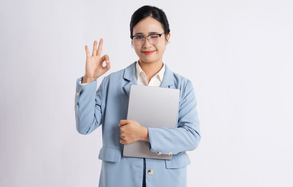 Mulher profissional de negócios com óculos, cabelo escuro, vestida formalmente com terno azul claro, sorrindo e fazendo sinal de aprovação com a mão, segurando um laptop e transmitindo confiança e otimismo.
