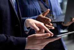 Profissionais de terno utilizando tablet e notebook em ambiente corporativo.