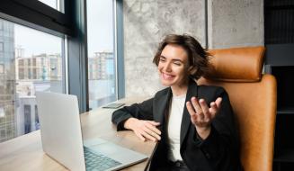 Mulher jovem e sorridente com cabelo curto em uma videochamada no laptop. Ela gesticula com a mão, veste blazer preto e está sentada em um escritório moderno com uma grande janela ao fundo.