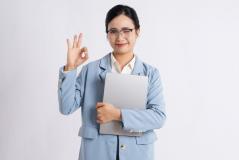 Mulher profissional de negócios com óculos, cabelo escuro, vestida formalmente com terno azul claro, sorrindo e fazendo sinal de aprovação com a mão, segurando um laptop e transmitindo confiança e otimismo.