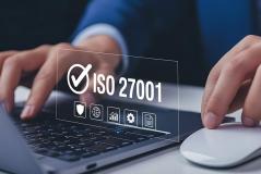 Close-up das mãos de uma pessoa usando um notebook e mouse, com uma sobreposição digital do selo de certificação ISO 27001 e ícones de segurança cibernética.