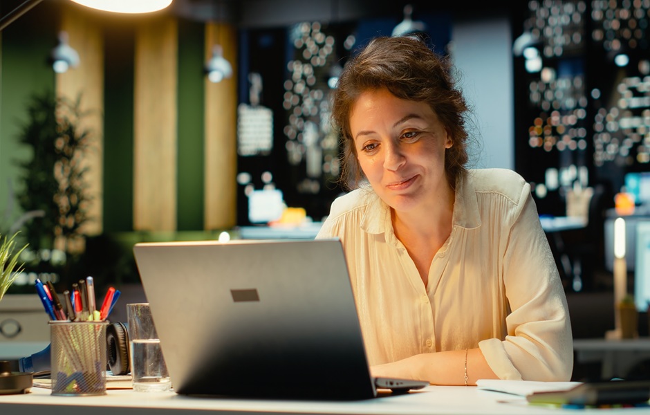 Mulher sorridente trabalhando em seu laptop em um ambiente moderno, com iluminação agradável e decoração contemporânea, ideal para produtividade.