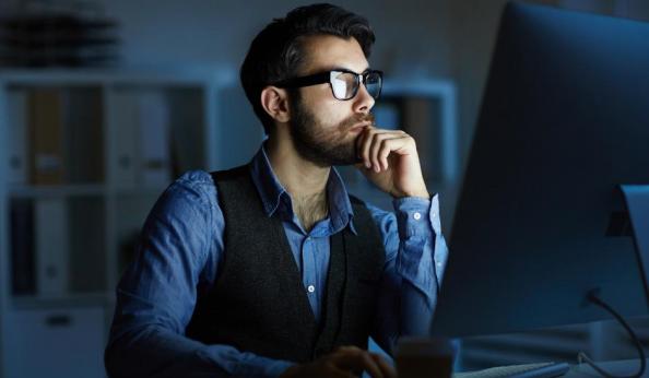 Homem barbudo de óculos e colete trabalhando concentrado em frente a um computador em um escritório escuro à noite.