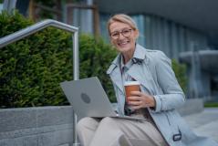 Mulher jovem e sorridente sentada ao ar livre com um laptop e um café, desfrutando do ambiente urbano e do clima agradável, transmitindo produtividade e bem-estar.