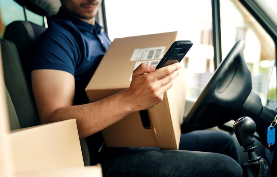 Entregador sentado no banco do motorista de uma van de entregas, segurando uma caixa de papelão com código de barras e verificando o celular. Ao fundo, outras caixas e o painel do veículo.