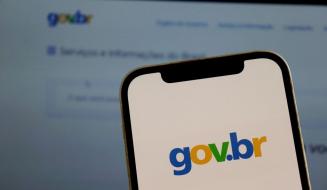 Imagem de um smartphone exibindo o logotipo do site gov.br com fundo borrado da tela de um computador, representando acesso ao portal oficial do governo brasileiro.