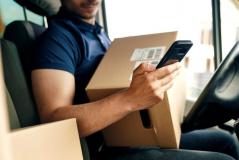 Entregador sentado no banco do motorista de uma van de entregas, segurando uma caixa de papelão com código de barras e verificando o celular. Ao fundo, outras caixas e o painel do veículo.